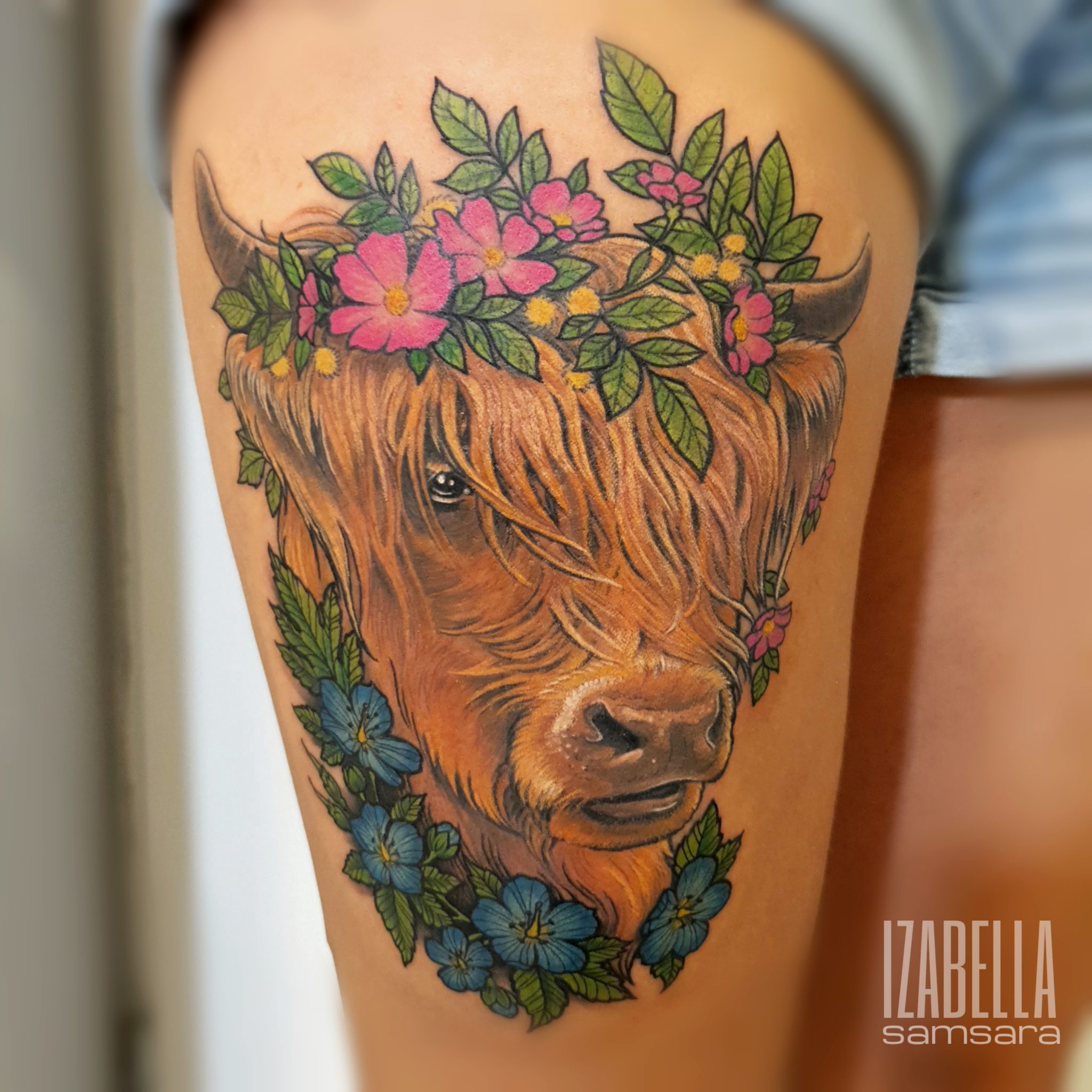A vibrant tattoo of a Highland cow's face, adorned with colorful flowers and green foliage, showcasing intricate details. The cow has a shaggy coat and expressive eyes, surrounded by pink and blue blossoms, adding a floral touch to the rustic animal design.