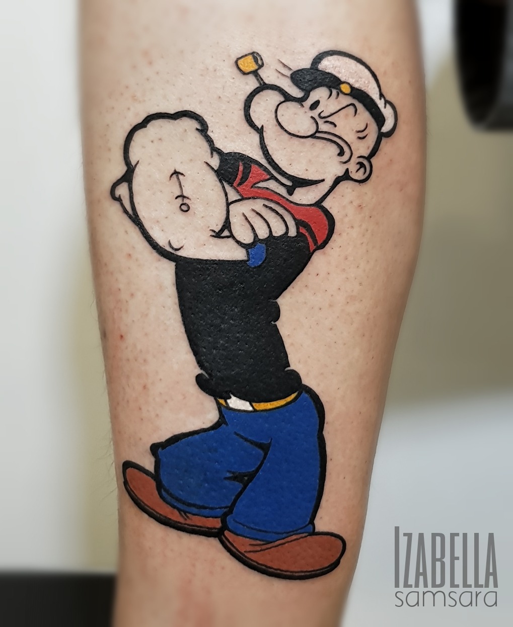Tattoo of Popeye the Sailor Man flexing his bicep, featuring vibrant colors including blue pants, a black shirt, and brown shoes, with his iconic sailor hat and pipe.