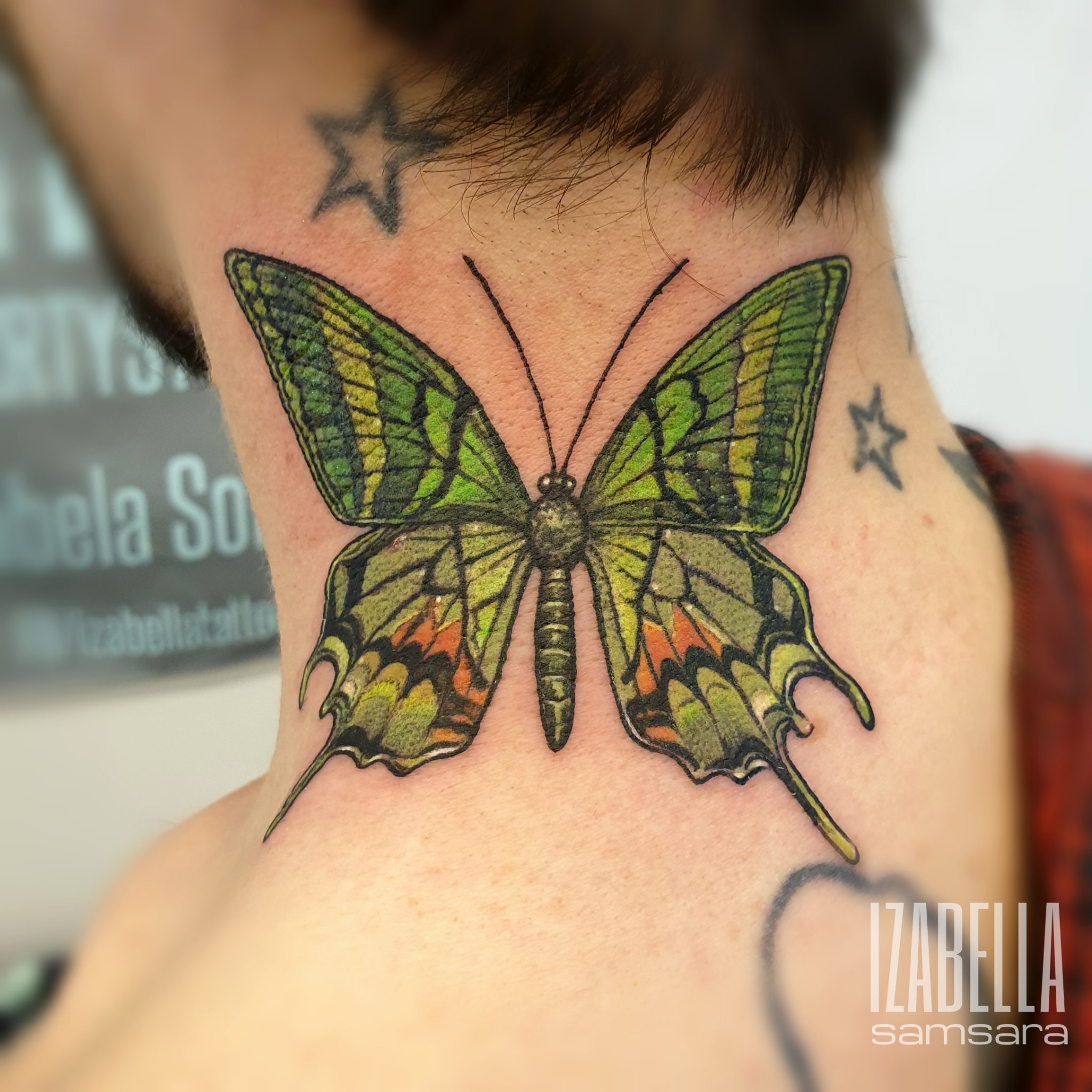 A vivid tattoo of a butterfly located on the nape of a neck, featuring a detailed design with green and orange hues. The wings are intricately illustrated, showcasing patterns and textures typical of a butterfly's anatomy, with a smooth gradient transitioning from green to orange near the edges. The tattoo is surrounded by a star tattoo in the background.