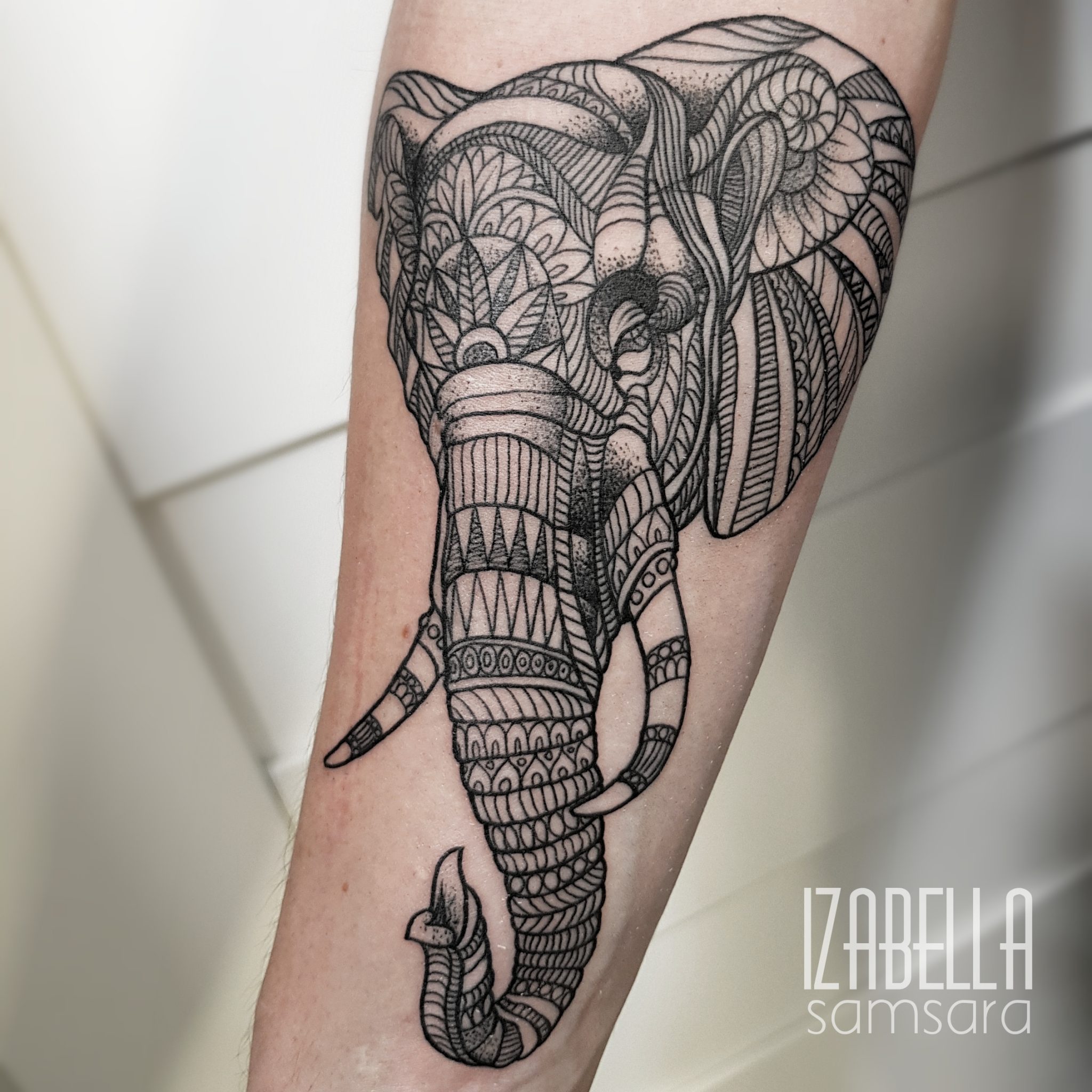 Detailed black and white tattoo of an elephant on the forearm, featuring intricate patterns, geometric designs, and stylized features, showcasing the elephant's trunk and ears. The tattoo emphasizes fine line work and shading, creating a harmonious yet captivating design.