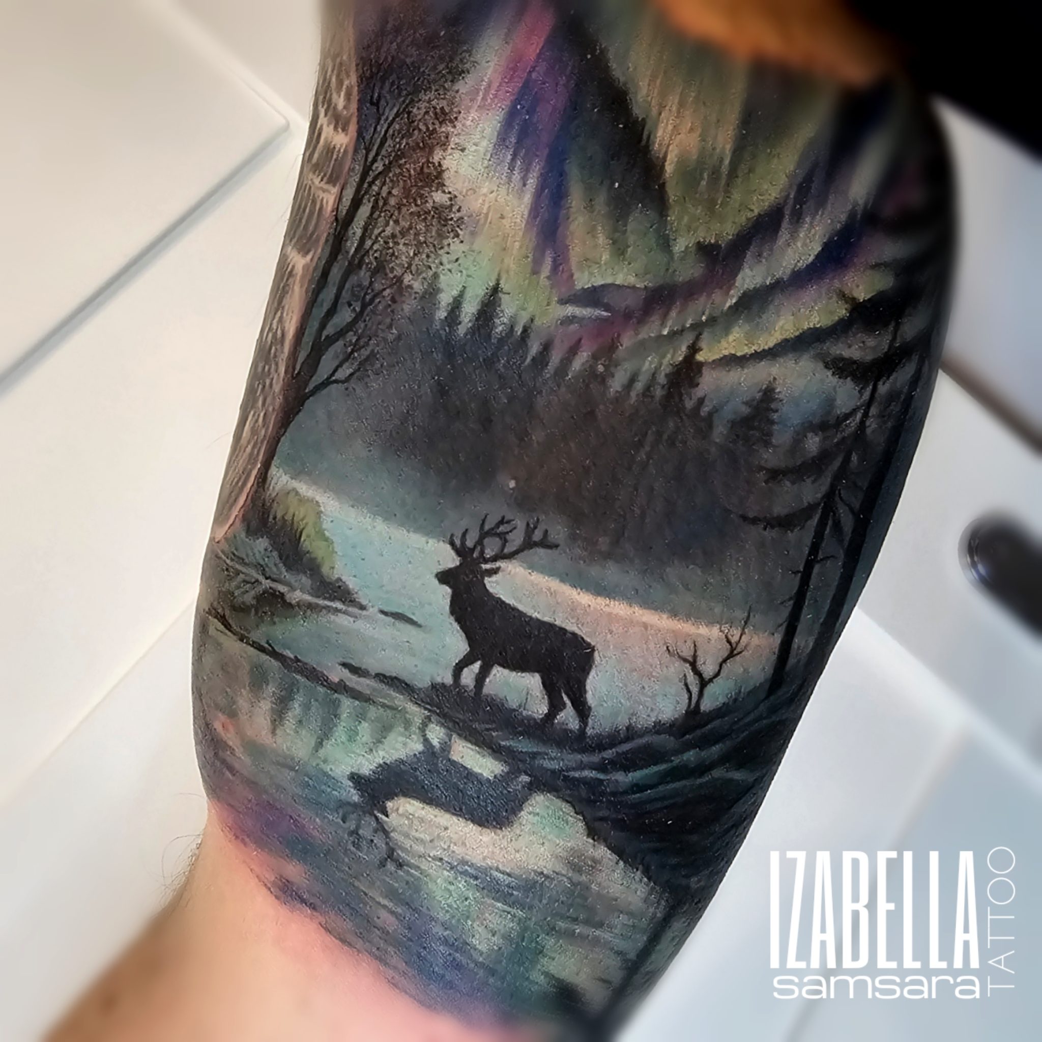 A beautifully detailed tattoo featuring a silhouette of a stag standing by a tranquil river with reflections in the water. The background showcases a scenic landscape with trees and a stunning aurora borealis illuminating the sky in vibrant colors. This tattoo combines elements of nature and wildlife, creating a serene and captivating design on the arm.