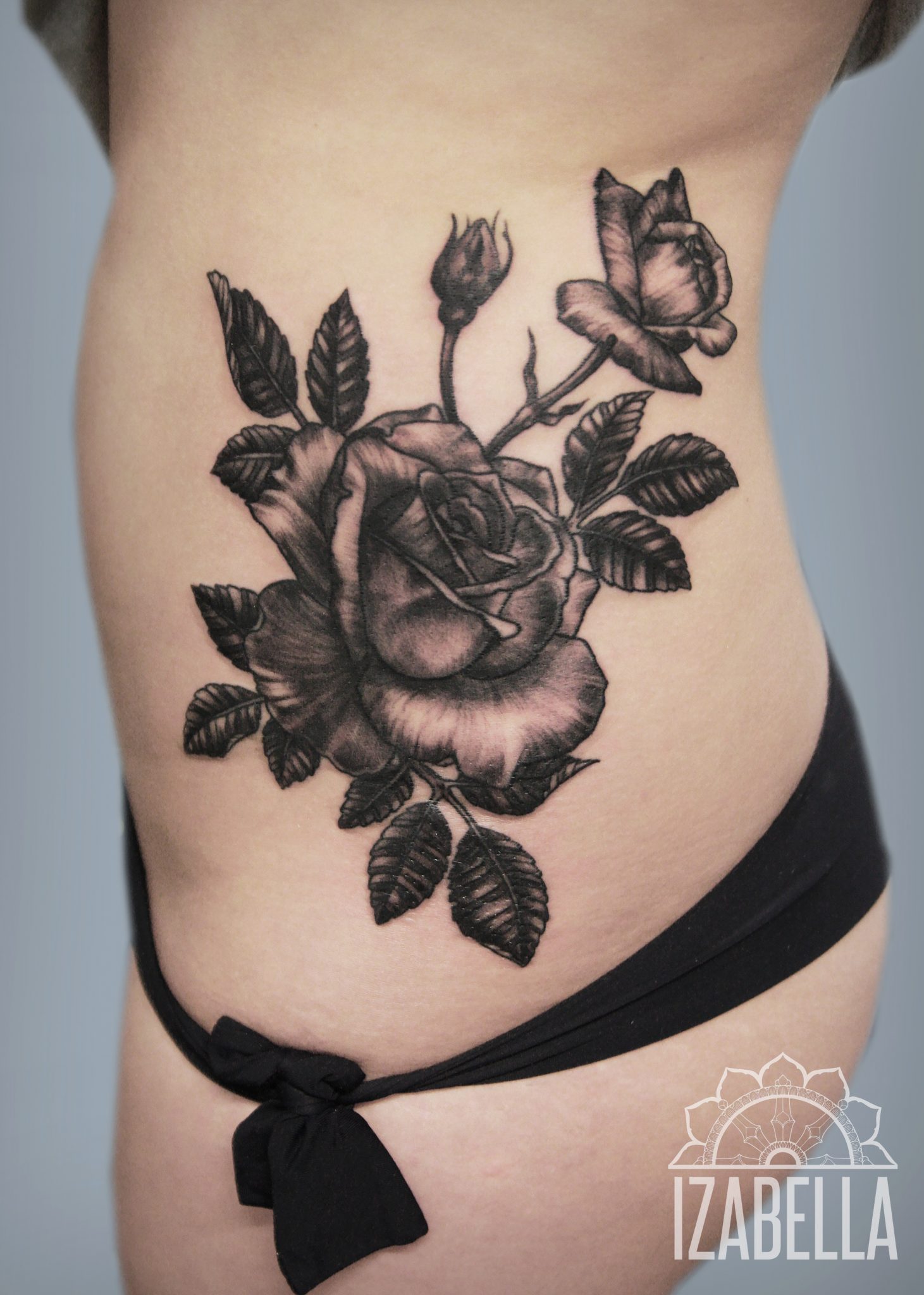 A black and gray tattoo of a large open rose and a smaller rosebud, surrounded by detailed leaves. The tattoo is placed on the side of the body, showcasing bold outlines and shading that create a realistic appearance.