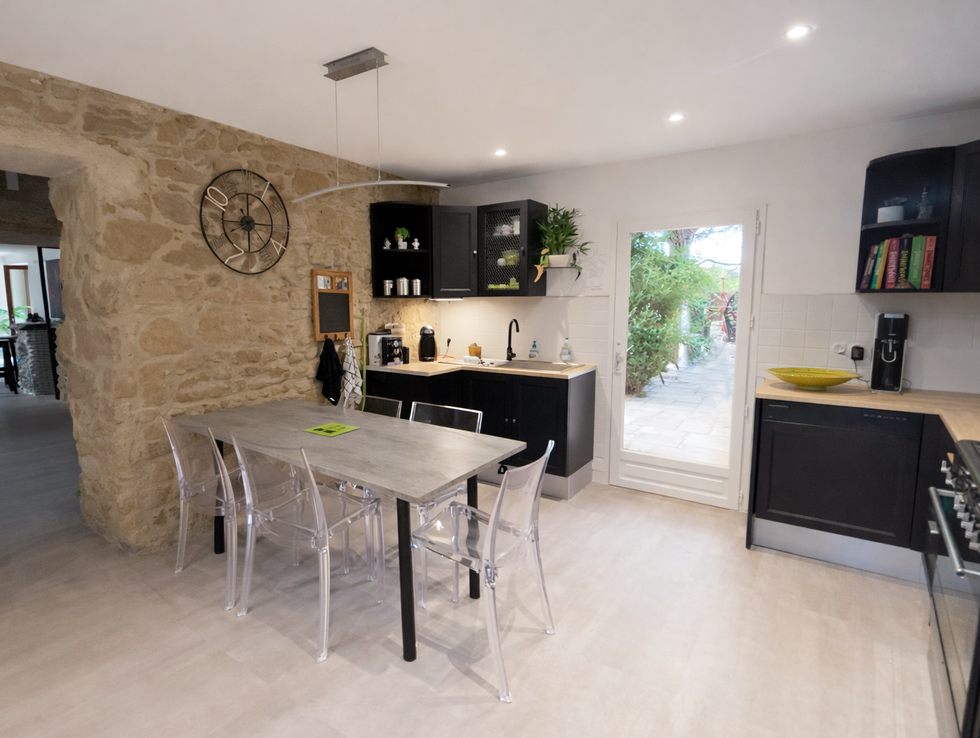 Cuisine avec mur en pierre apparente, meubles noirs et bois clair, table rectangulaire avec chaises transparentes et porte vitrée ouvrant sur l’extérieur.