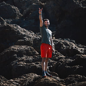 Fotograf steht auf dunklen Felsen im Freien, lächelt in die Kamera und zeigt ein Peace-Zeichen mit erhobenem Arm.