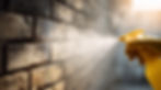 Gloved hand sprays clear liquid from yellow bottle onto a sunlit brick wall, creating a mist. Warm tones and soft, focused lighting.