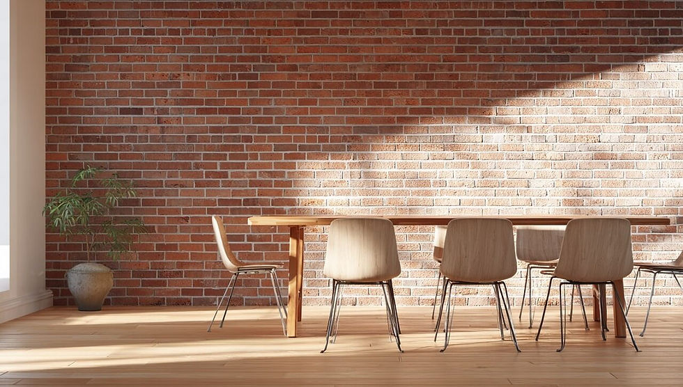 Dining table with six wooden chairs on a sunlit wooden floor, set against a red brick wall. A potted plant stands on the left.
