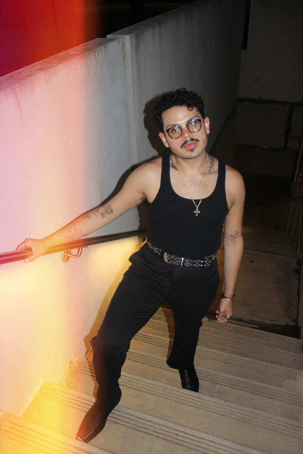 Person in a black tank top and glasses stands on stairs, holding a railing. Tattoos visible. Warm light leak effect on left side.