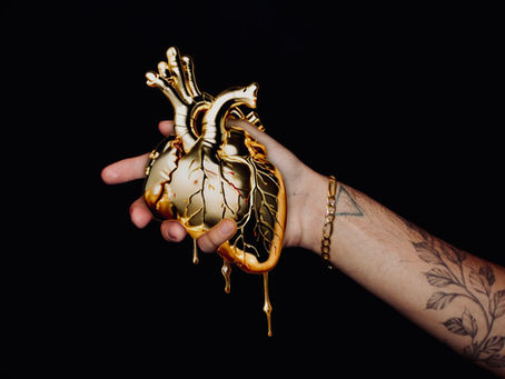 Tattooed hand holds a dripping golden heart against a black background. The heart's metallic sheen contrasts with the dark setting.