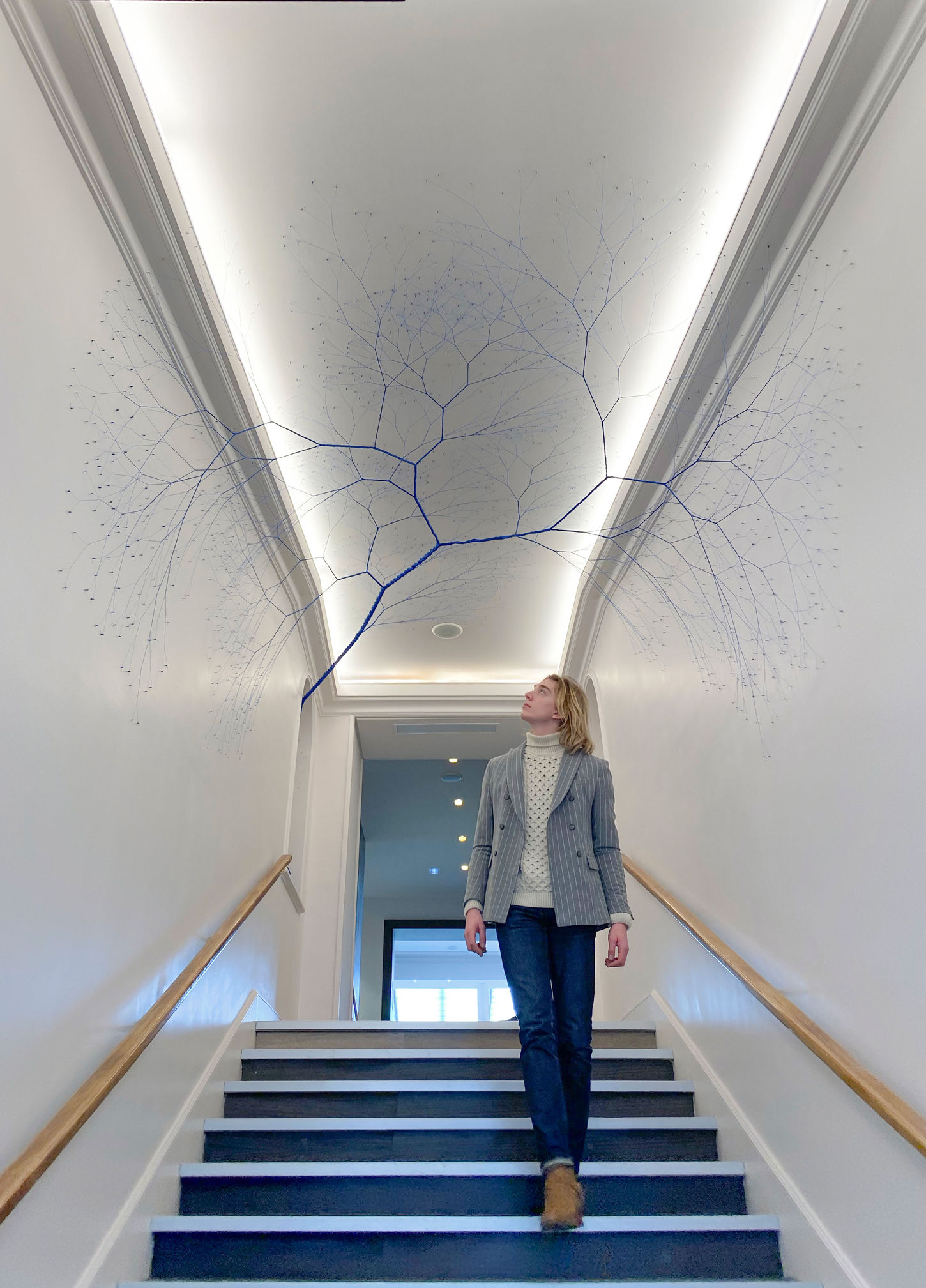 Site-specific installation titled "Ciclotrama 175 (impregação)" by Janaina Mello Landini in Cannes, France. The sculpture begins as a thick coil of bright blue rope on a white ledge beside a flight of indoor stairs. The rope unwinds and stretches vertically upward before branching out into a complex, fine, dendritic web of individual blue threads that sprawl across the white ceiling and walls like vascular systems or root networks.