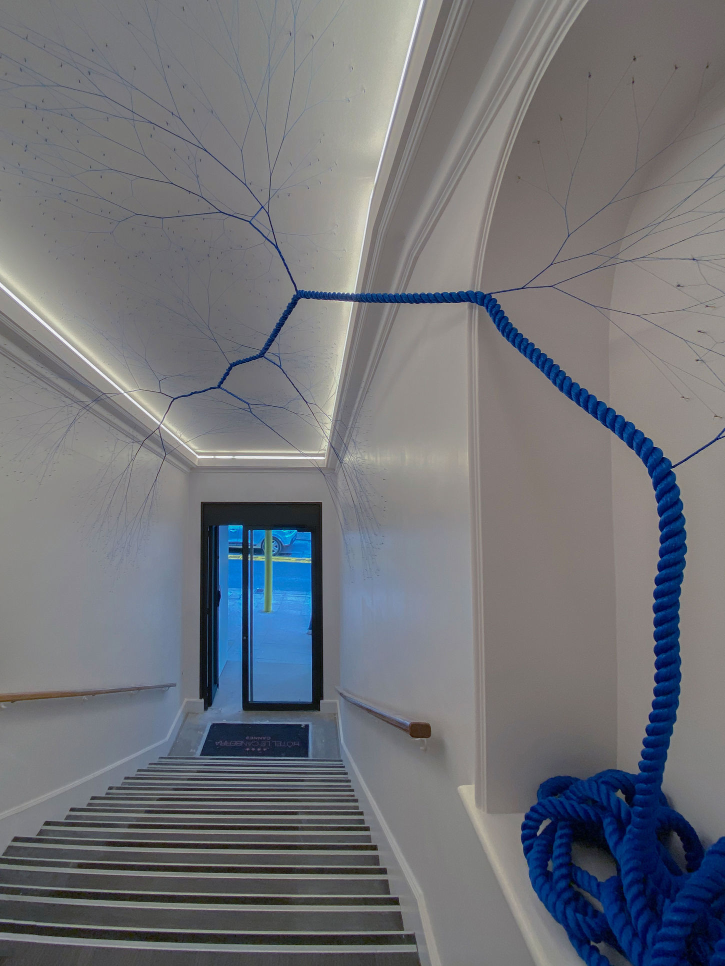 Site-specific installation titled "Ciclotrama 175 (impregação)" by Janaina Mello Landini in Cannes, France. The sculpture begins as a thick coil of bright blue rope on a white ledge beside a flight of indoor stairs. The rope unwinds and stretches vertically upward before branching out into a complex, fine, dendritic web of individual blue threads that sprawl across the white ceiling and walls like vascular systems or root networks.