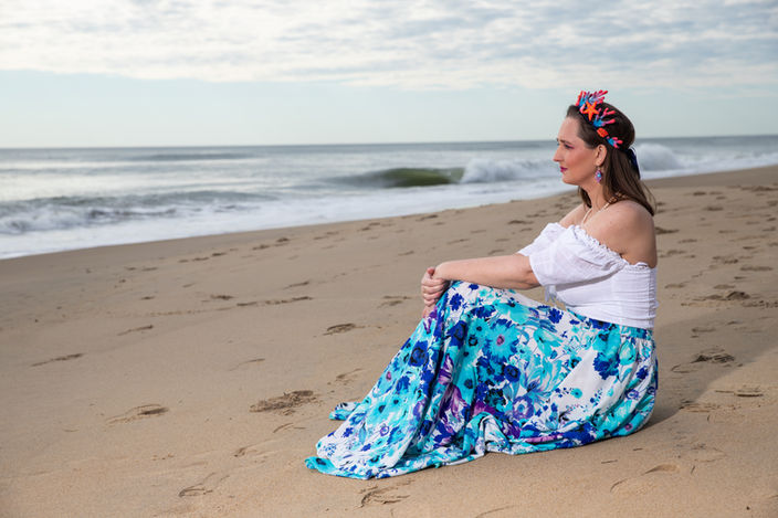 Woman sitting on beach, looking towards the ocean, wearing floral skirt and top.