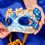 Thumbnail: Woman holding a decorative blue and white mask above blue fabric background.