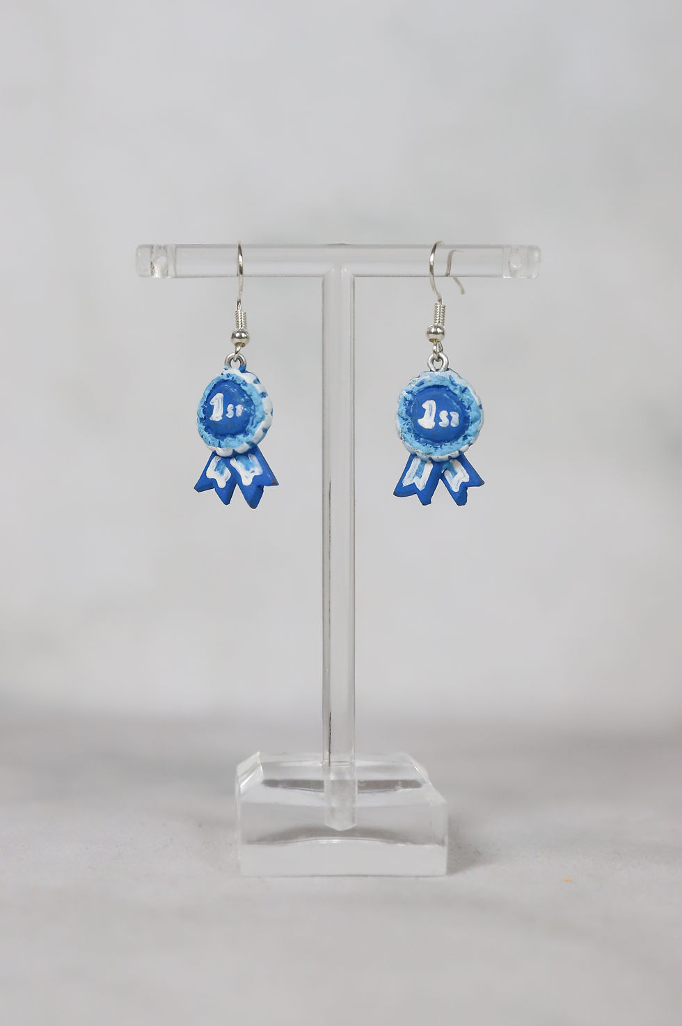 Pair of blue and white '1st' ribbon earrings displayed on a stand.