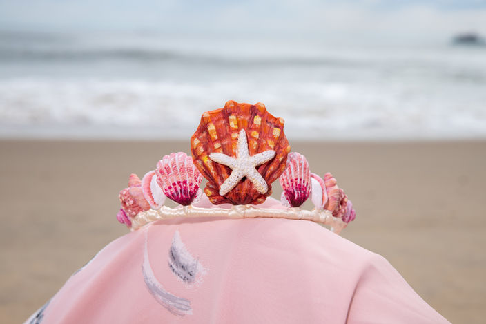 Decorative seashell crown on a person's head with ocean backdrop.