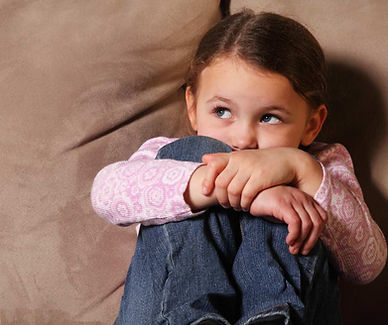 Young girl sits on a beige couch, hugging her knees, wearing a pink patterned shirt and jeans. She looks to the side, appearing pensive.