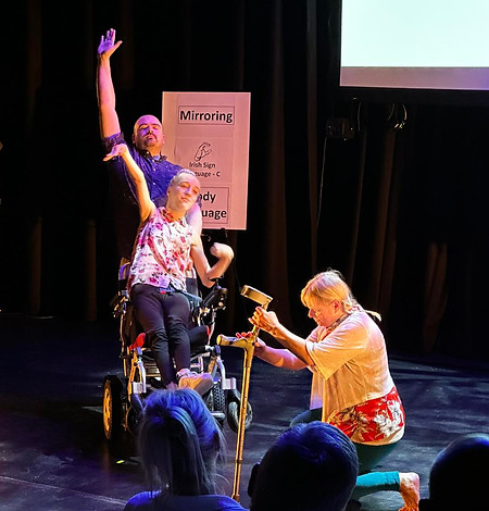 Talia is on stage dancing with two standing dancers. There is a projection on the screen behing them of a pair of human eyes staring out at the audience. Three display boards list information about different types of communication, such as mirroring, symbols and eye contact.