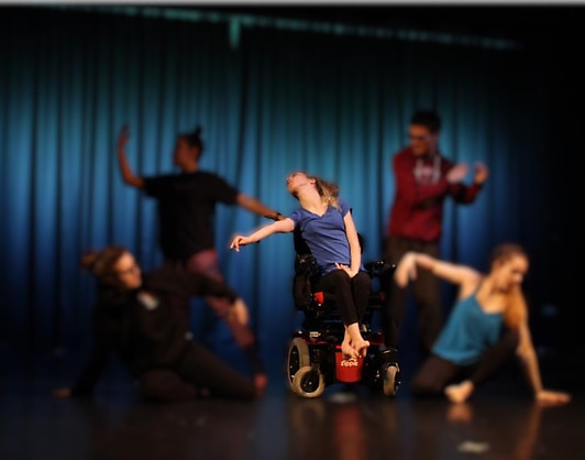 Talia dancing on stage in a purple top and black leggings. Her blonde hair is tied in a ponytail, she is seated in her power chair and her feet are bare. Other dancers are on the stage in the background but their images are blurred out.