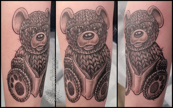 tattoo of a knitted, teddy bear, toy, reading a book and wearing glasses.  sharptattoos