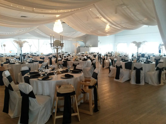 An indoor wedding reception set up by Chef Mike and his team with round tables covered in white table clothes with black runners and white cloth chairs with black ribbons