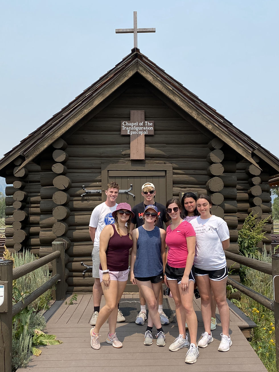 The seven young pilgrims stand in front of a chapel, built in a log cabin style.