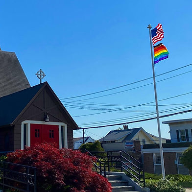 In front of a blue sky, the entrance to St. Paul's stands next to a large flag pole with an American flag and a Philadelphia LGBTQIA+ pride flag underneath