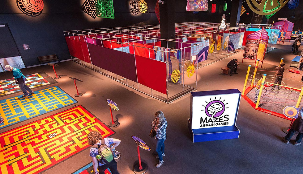 "Visitors exploring interactive floor mazes and colorful exhibits at the 'Mazes & Brain Games' event, with a prominent logo stand in the foreground."