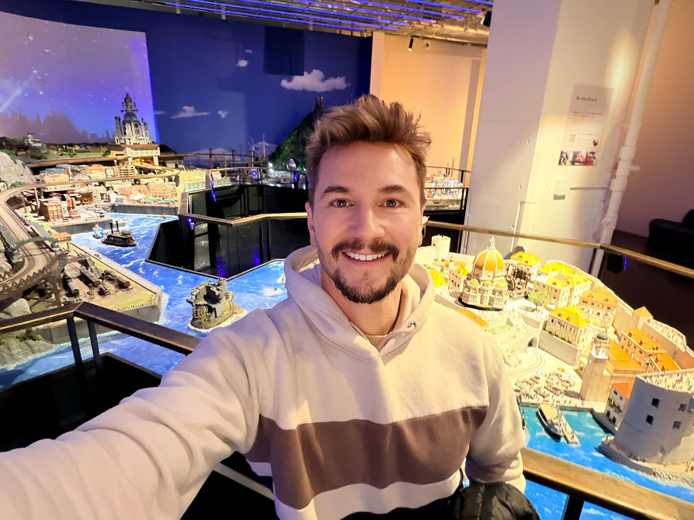 Smiling person takes a selfie in front of a detailed miniature cityscape with blue water and colorful buildings in a brightly lit exhibit.