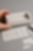 Hand holds a miniature paper sofa model next to sketches and paper pieces on a gray surface. Tweezers are visible, creating a crafty, detailed mood.