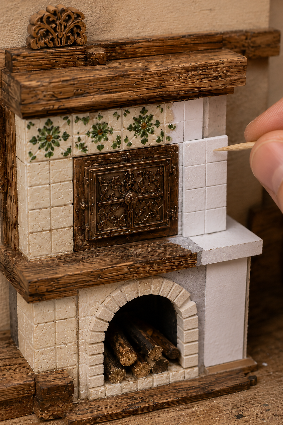 Hand painting a miniature ornate tiled stove model with a toothpick, showing carved wood, floral tiles, and stacked logs.