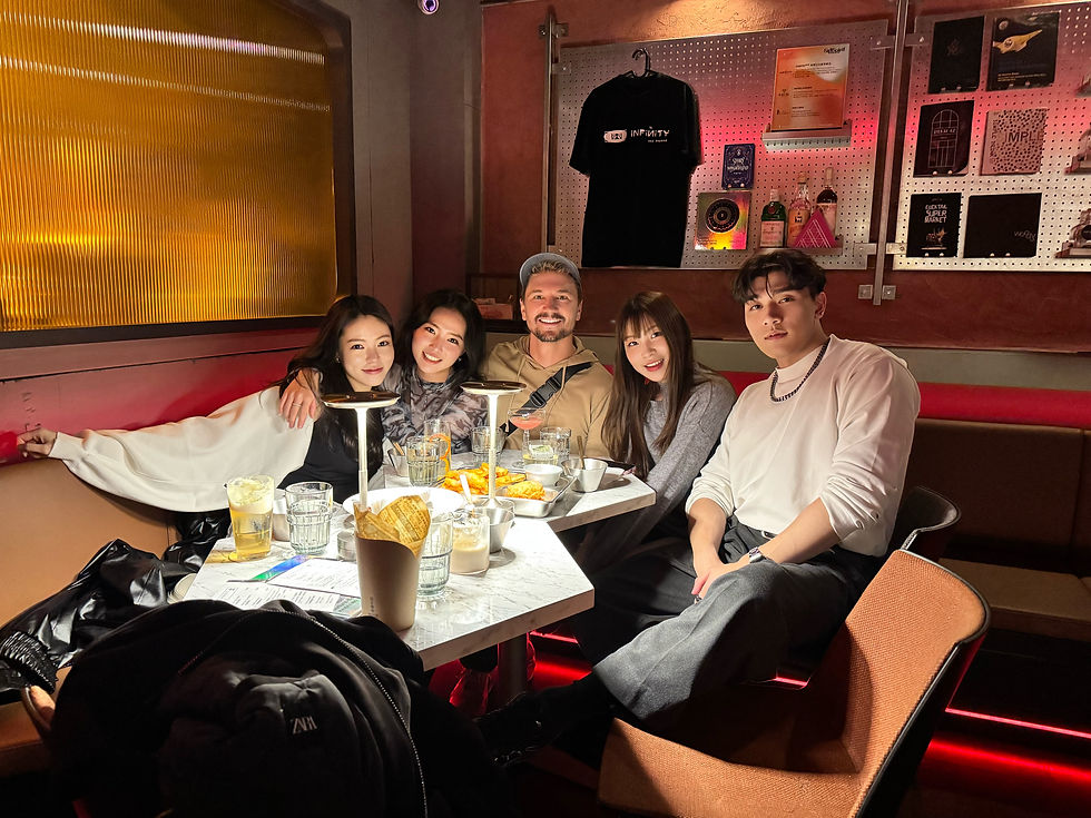A group of five friends sits closely at a restaurant table with drinks and snacks. The scene is warmly lit, and a "ZARA" jacket is visible.