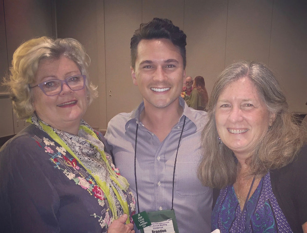 Three people smiling at a conference, wearing name badges. The woman on the left wears glasses and a floral scarf. Neutral-colored background.