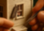 Hands use tweezers to adjust a small window shutter on a detailed model house. A striped curtain is visible inside. Mood is focused.