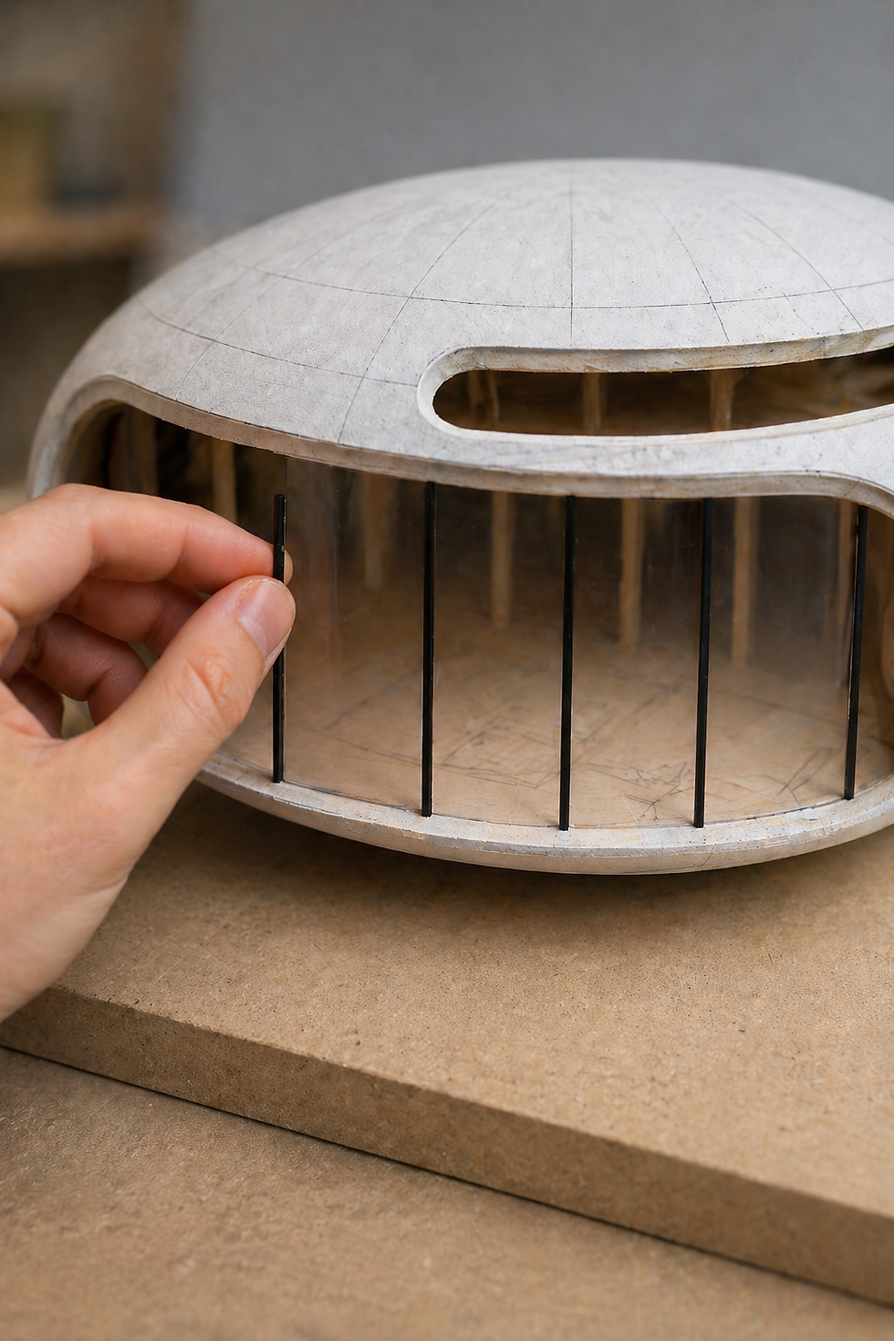 A hand places a black stick into a futuristic, dome-shaped architectural model with clear panels on a beige surface.