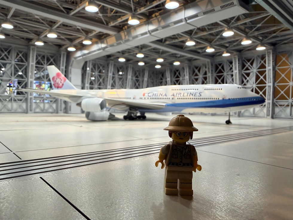 Lego figure stands in a hangar with a China Airlines plane in the background. Bright lights above; the plane's tail features a floral design.