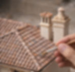 Hand painting a miniature house roof with a brush. Earth-tone paint palette visible. The setting conveys a delicate, artistic mood.