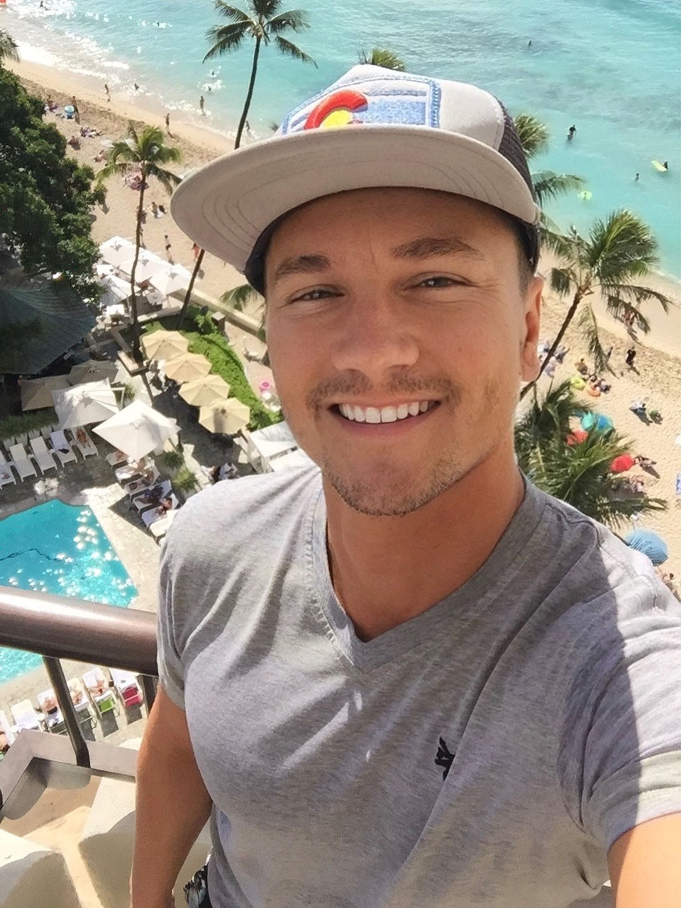Man in a gray shirt and cap smiles for a selfie on a balcony overlooking a beach and ocean. Palms and clear blue water in the background.