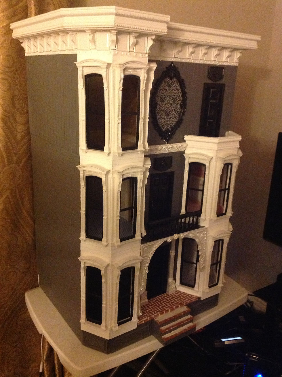 Victorian-style dollhouse with intricate white trim, black windows, and a brick front step, set against a patterned curtain background.