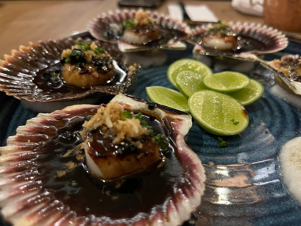 Scallops in shells with sauce and garnish on a blue plate, alongside sliced limes. Warm, upscale dining setting.