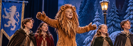 Young performers on a snowy Narnia stage with a child dressed as a lion beside a glowing lamppost.