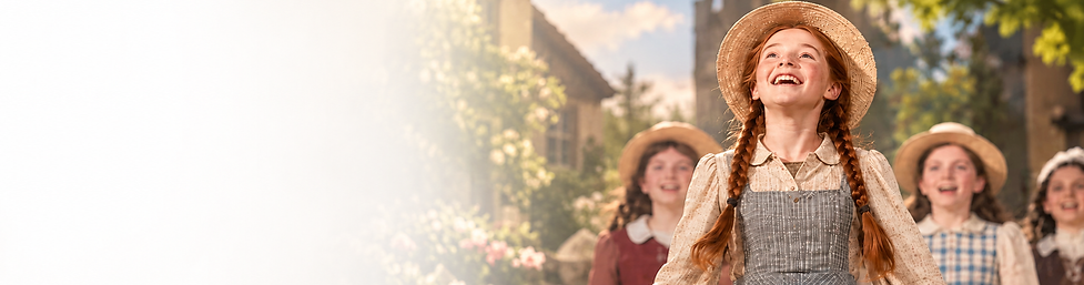 Young performer dressed as Anne of Green Gables, with red braids and a straw hat, smiling on stage with other children.