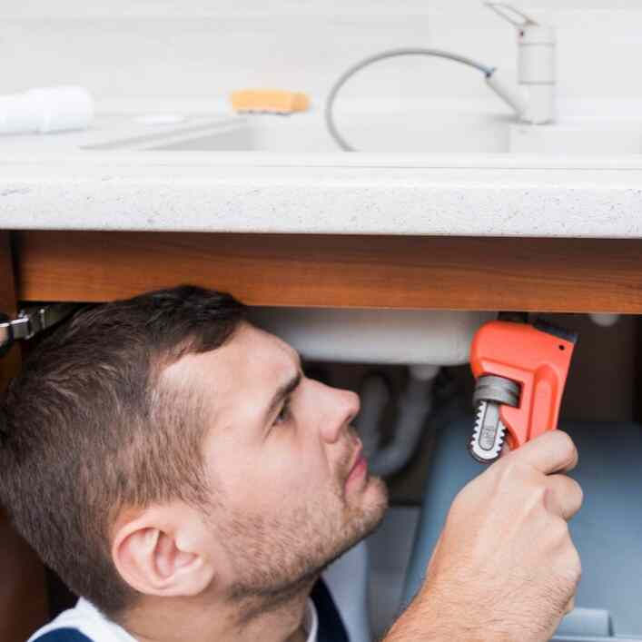 Man fixing a pipe under a kitchen sink with a red wrench. Focused expression, countertop and faucet in background, bright lighting.