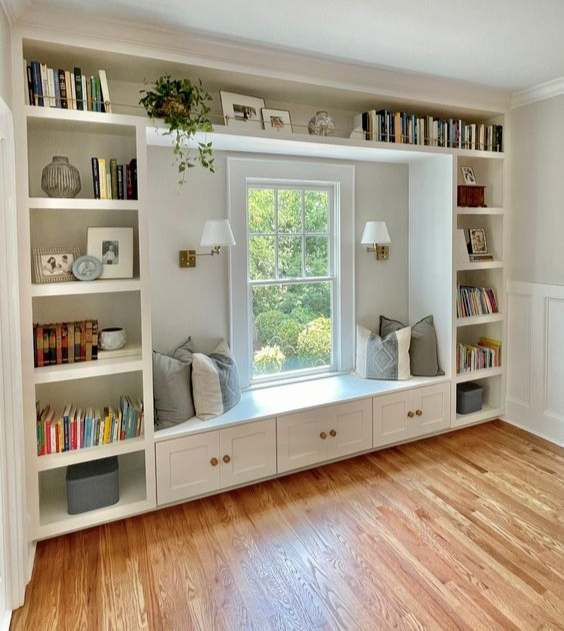 Built-in bookshelves surround a window seat with gray cushions. Books, plants, and decor adorn the shelves. Light wood floor, bright ambiance.