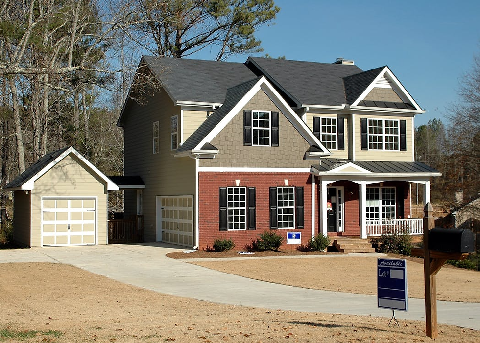 Two-story house with brick and beige siding, black shutters, detached garage, for sale sign in yard, driveway, trees in background, sunny day.