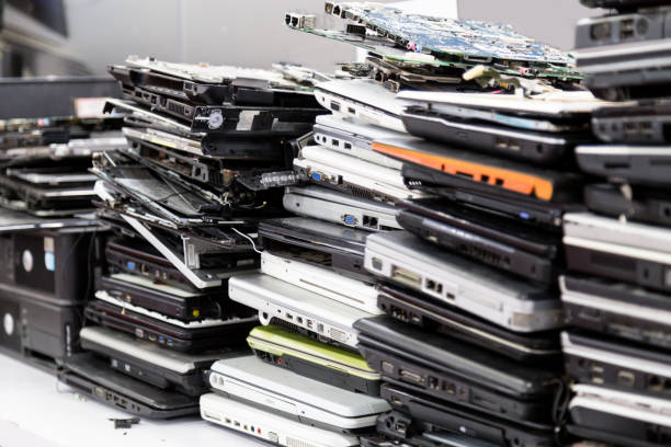 Stacks of old, variously colored laptops piled in disarray indoors, suggest a sense of clutter and e-waste.