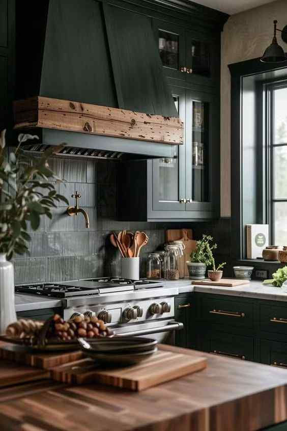 Cozy kitchen with dark green cabinets, wooden accents, and black counters. Stove, utensils, potted herbs, and fruit on a wooden counter. Natural light.