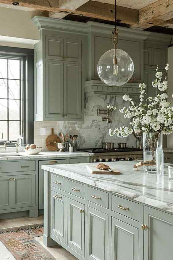Elegant kitchen with sage green cabinets, marble countertops, and a large pendant light. A vase with white flowers adds a fresh touch.