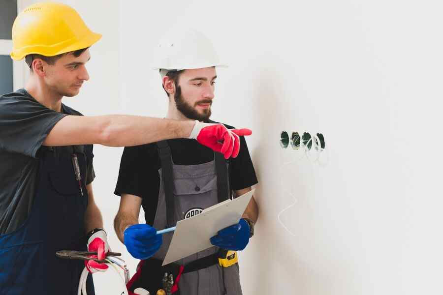 Two construction workers in hard hats and gloves examine wall wiring. One points while the other holds tools and a clipboard in a bright room.