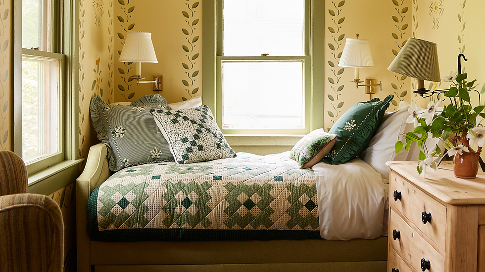Cozy bedroom with floral wallpaper, green quilted bed, and pillows. Wooden dresser with flowers in vase. Sunlight through windows.