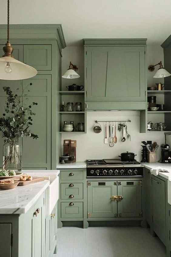 Cozy kitchen with sage green cabinets, marble countertop, and vintage stove. Hanging utensils, plants in vase, and warm lighting.