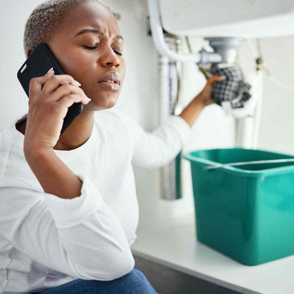 A person in a white shirt calls on their phone, holding a pipe under a sink. A green bucket is placed to catch water. They appear stressed.